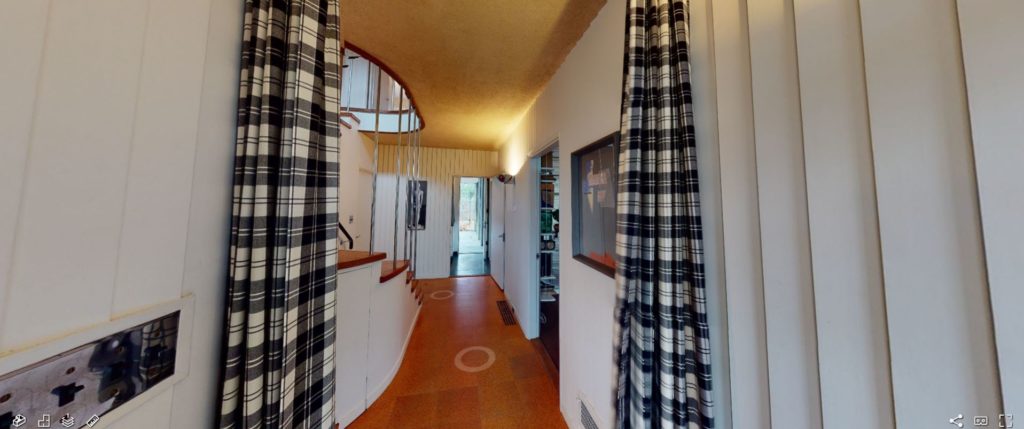 A narrow interior hallway with hard wooden floors and a white paneled wall. There is a small wooden staircase on the left side and an entry way to a room to the right.